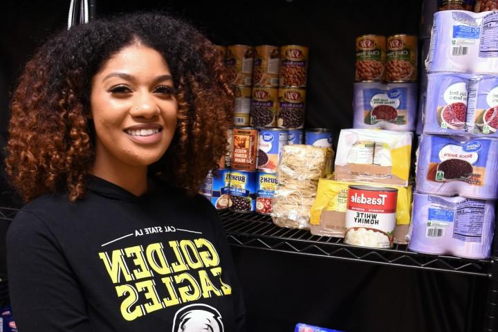 A person smiling, wearing a Golden Eagles sweatshirt, standing in front of shelves stocked with non perishable food.